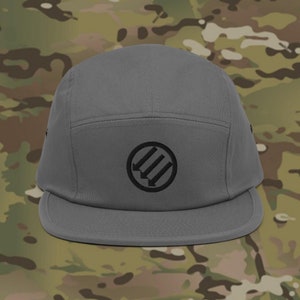 May include: A gray five-panel cap with a flat brim. The cap features a black circular emblem with three downward-pointing arrows. The hat is made of a durable fabric and is suitable for outdoor wear. The background is a camouflage pattern.