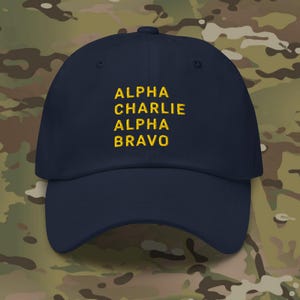 May include: A navy blue baseball cap with yellow embroidered text that reads "ALPHA CHARLIE ALPHA BRAVO".