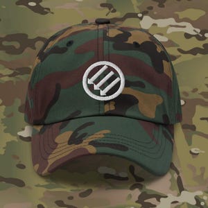 Puede incluir: Una gorra de béisbol de camuflaje con un círculo bordado blanco que contiene tres flechas blancas que apuntan hacia arriba.