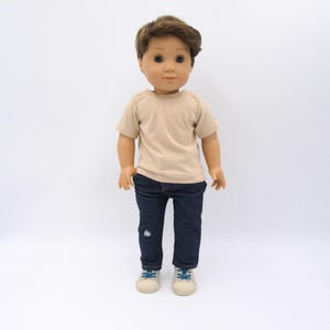 K&ouml;nnte beinhalten: Eine Puppe mit braunen Haaren und braunen Augen, bekleidet mit einem beigen T-Shirt mit kurzen &Auml;rmeln, einer dunkelblauen Jeans mit einem kleinen wei&szlig;en Stickdetail am linken Bein und hellen Turnschuhen mit blauen Schn&uuml;rsenkeln vor wei&szlig;em Hintergrund.