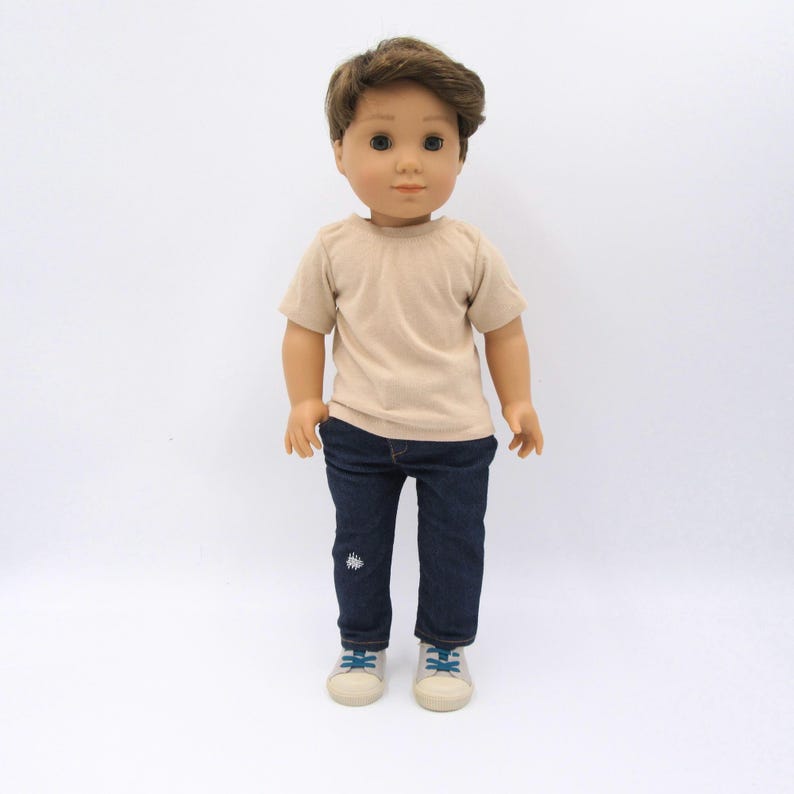 K&ouml;nnte beinhalten: Eine Puppe mit braunen Haaren und braunen Augen, bekleidet mit einem beigen T-Shirt mit kurzen &Auml;rmeln, einer dunkelblauen Jeans mit einem kleinen wei&szlig;en Stickdetail am linken Bein und hellen Turnschuhen mit blauen Schn&uuml;rsenkeln vor wei&szlig;em Hintergrund.