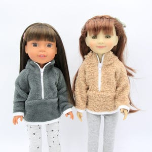 May include: Two dolls wearing fleece zip-up sweaters. The doll on the left is wearing a gray sweater with a white zipper and white trim. The doll on the right is wearing a beige sweater with a white zipper and white trim. Both dolls are wearing gray leggings.