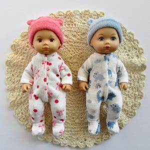 May include: Two baby dolls dressed in matching sleepsuits and hats. One doll wears a pink hat and cherry-patterned pyjamas. The other has a blue hat and dinosaur-patterned sleepsuit. They are on a cream-coloured, crocheted doily.