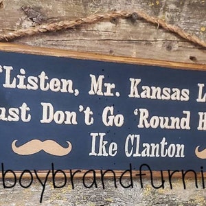 May include: A dark blue wooden sign with white text that reads "Listen, Mr. Kansas Law Dog, Law Just Don't Go 'Round Here. Savvy?"  The sign also includes the name "Ike Clanton" and a hashtag that reads "#cowboybrandfurniture".