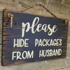 May include: A rectangular wooden sign with the words "please HIDE PACKAGES FROM HUSBAND" in white and yellow lettering. The sign is black with a distressed finish and is suspended by a rope.