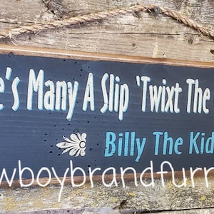 Könnte beinhalten: Rechteckiges Holzschild mit dunkelblauem Hintergrund und weißem Text, der lautet: "There's Many A Slip 'Twixt The Cup And The Lip!" und "Billy The Kid". Das Schild ist braun eingefasst und hat eine Seilaufhängung.
