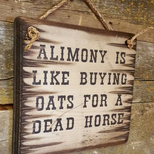 May include: A distressed wooden sign with the phrase "ALIMONY IS LIKE BUYING OATS FOR A DEAD HORSE" in black. The sign has a dark brown edge and is hung by a natural rope.