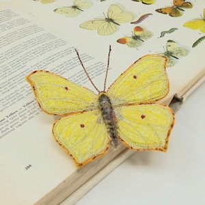 May include: A yellow butterfly with brown markings is displayed on an open book. The butterfly is made of fabric and has a delicate, embroidered texture.