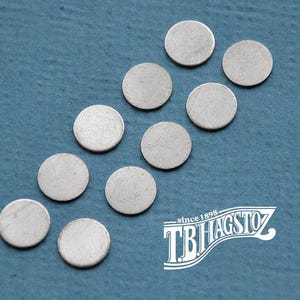 May include: Ten small, circular, flat metal discs arranged in a diagonal pattern. Each disc has a smooth, matte silver-colored surface. The white logo text TB HAGSTOZ since 1898 is positioned in the lower right corner of the frame.