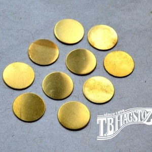 May include: Ten small, flat, circular gold-colored metal discs arranged in a loose cluster. Each disc has a smooth, reflective surface. In the bottom right corner, there is a white logo with the text "since 1898 TBHAGSTOZ" in a stylized font.