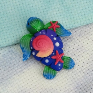 May include: A colorful polymer clay turtle figurine. The turtle has a blue shell with white dots, a spiral design, and red starfish accents. Its flippers and head are green and blue. The figurine is on a light blue and white patterned surface.
