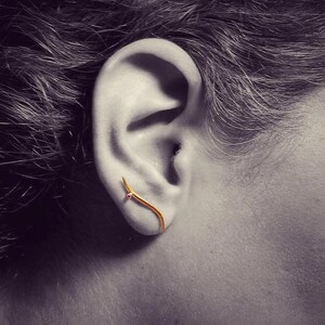 Könnte beinhalten: Ein goldener Ohrring mit einem kleinen rosafarbenen Edelstein. Der Ohrring ist gebogen und hat ein spitzes Ende.