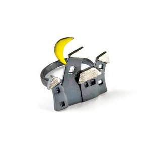 Puede incluir: Un anillo de plata con un grupo de tres pequeñas casas y una luna creciente amarilla. Las casas son grises con techos blancos.