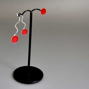 May include: A pair of modern earrings on a black display stand. The earrings feature a silver, wavy design with a small, round, red accent. The display stand is black with a circular base and a vertical post.