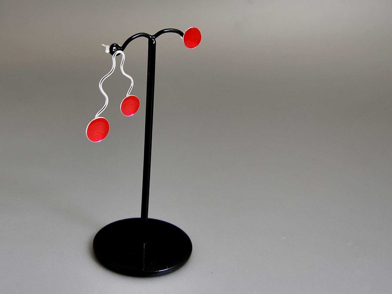 May include: A pair of modern earrings on a black display stand. The earrings feature a silver, wavy design with a small, round, red accent. The display stand is black with a circular base and a vertical post.