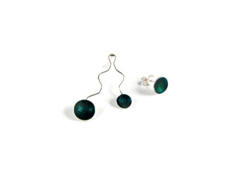 May include: A jewelry set featuring a pendant and stud earrings. The pendant has two dark teal, glittery circular pendants connected by a silver, wavy wire. The stud earrings have a similar design. All pieces are set against a white background.