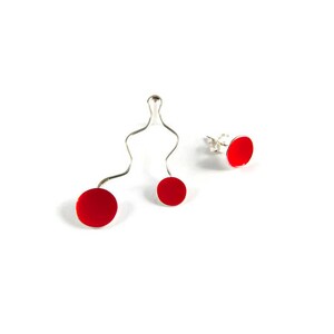 May include: A set of three pieces of jewelry: a pair of red stud earrings and a silver wire pendant with two red circular accents. The jewelry is set against a white background.