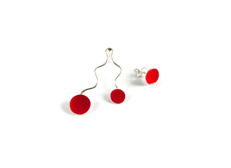 May include: A set of three pieces of jewelry: a pair of red stud earrings and a silver wire pendant with two red circular accents. The jewelry is set against a white background.