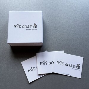 May include: A white square box and three white cards with the text "this and that" in a handwritten style, with a heart and crown graphic. The box and cards are on a gray surface.
