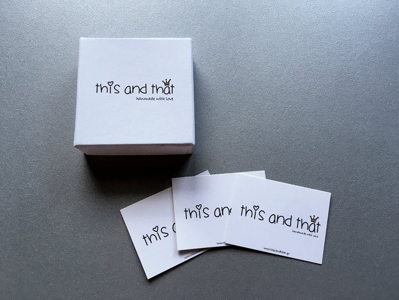 May include: A white square box and three white cards with the text "this and that" in a handwritten style, with a heart and crown graphic. The box and cards are on a gray surface.