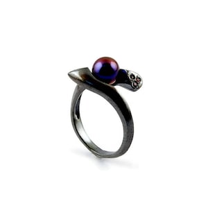 Könnte beinhalten: Ein silberner Ring mit einer schwarzen und violetten Perle und einem kleinen Cluster aus roten Edelsteinen.