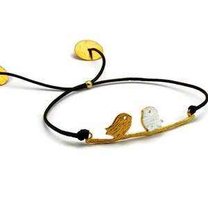 Op de afbeelding: Een gouden armband met twee vogels die op een tak zitten. De armband is verstelbaar met twee gouden schijven en een zwart koord.