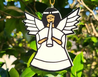 May include: A white angel ornament with black and brown details, hanging from a gold string. The angel is playing a horn, with outstretched wings. The ornament is hanging in front of green leaves. The tag reads "El Salvador".