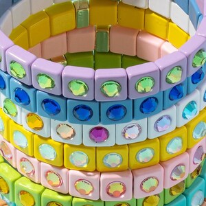 May include: Stack of colorful square-shaped bracelets with iridescent rhinestones. Each bracelet is made of different colored plastic squares with a single rhinestone in the center of each square.