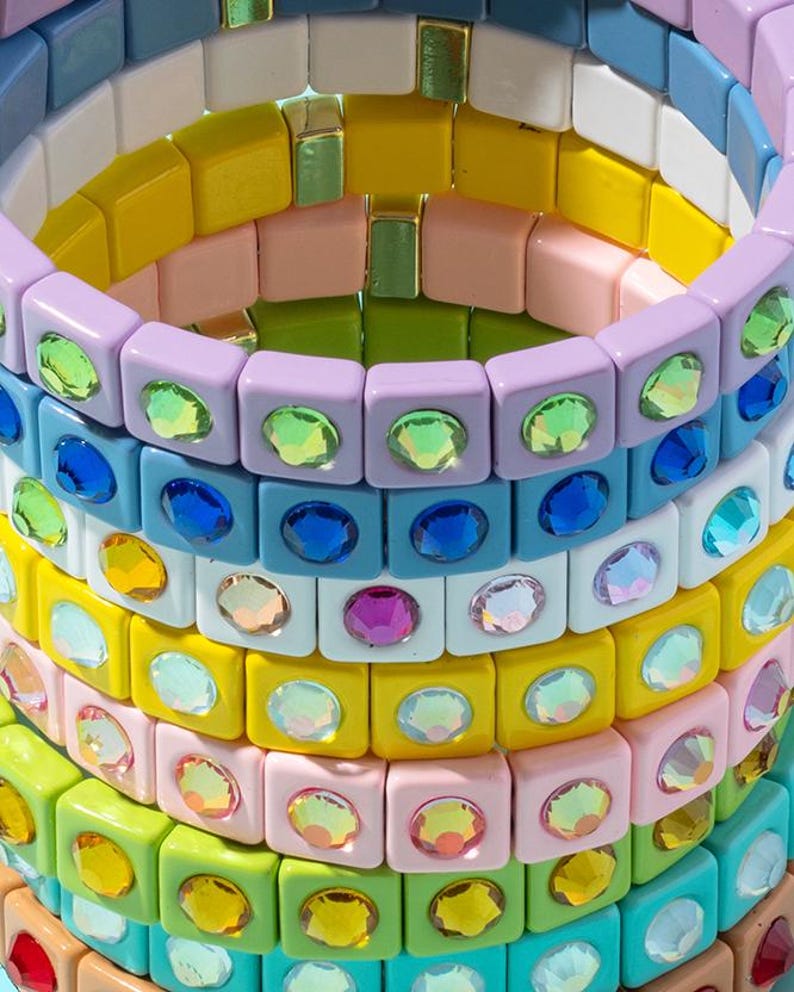May include: Stack of colorful square-shaped bracelets with iridescent rhinestones. Each bracelet is made of different colored plastic squares with a single rhinestone in the center of each square.