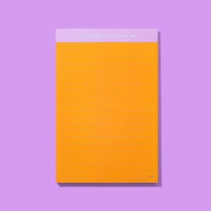 May include: A lined notepad with an orange paper and a lavender top. The text "be so good they can't ignore you" is printed in white on the lavender top. A small heart is in the bottom right corner. The notepad is on a purple background.