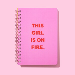 Op de afbeelding: Een roze spiraalgebonden notitieboek met de tekst "THIS GIRL IS ON FIRE" in het rood. Het notitieboek heeft een gele rug en een roze kaft. De achtergrond is effen roze.