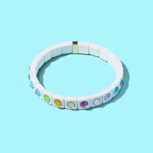 May include: White bracelet with colorful gemstones. The bracelet is made of square ceramic beads with a gold accent.