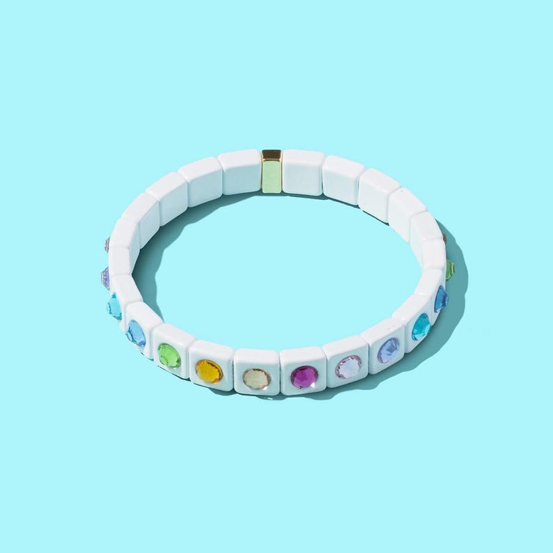 May include: White bracelet with colorful gemstones. The bracelet is made of square ceramic beads with a gold accent.