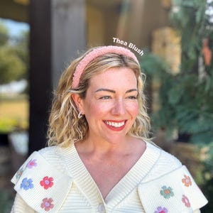 May include: A woman wearing a pink headband with the text "Thea Blush". She is wearing a cream-colored sweater with floral embroidery. The background includes greenery and a building.