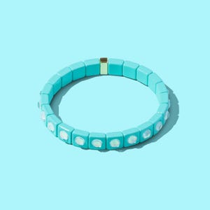 May include: A turquoise blue bracelet with white opal-like stones. The bracelet is made of square-shaped beads and has a gold-colored clasp.
