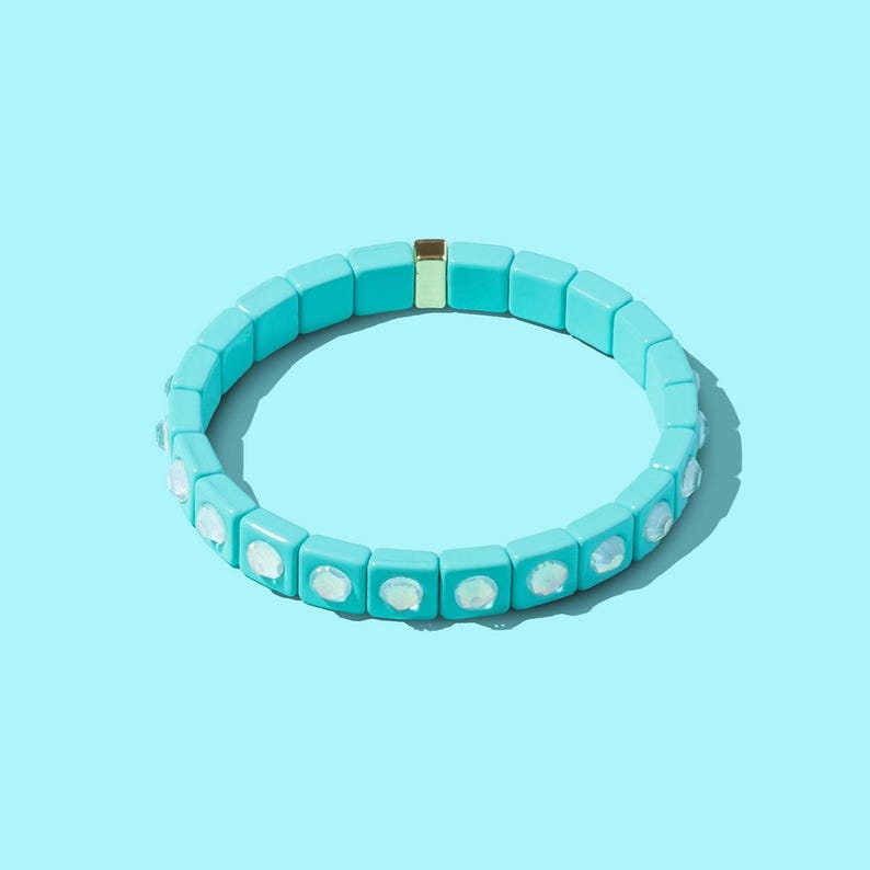 May include: A turquoise blue bracelet with white opal-like stones. The bracelet is made of square-shaped beads and has a gold-colored clasp.