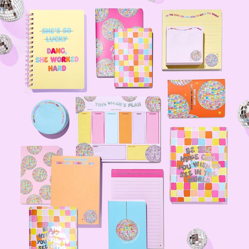 Op de afbeelding: Een verzameling kleurrijke schrijfwaren, waaronder notitieboekjes, notitieblokken en plaknotities, met disco-bal en geometrische ontwerpen. Het spiraalgebonden notitieboek heeft de tekst "SHE'S SO LUCKY, DANG, SHE WORKED HARD". Andere items bevatten de tekst "BE THE HOPE YOU WISH TO SEE IN THE WORLD".