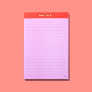 Op de afbeelding: Een roze gelijnd notitieblok met een rode header. De header heeft de gouden tekst "this girl is on fire". Het notitieblok heeft horizontale lijnen en een klein hartje onderaan.