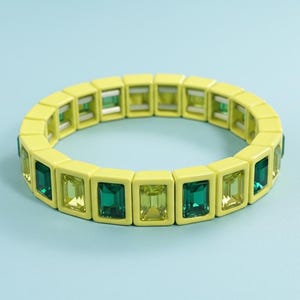 May include: A yellow bracelet with rectangular green and yellow gemstones. The bracelet is made of small, square yellow blocks, each holding a gemstone. The gemstones alternate between shades of green and yellow. The bracelet is set against a light blue background.