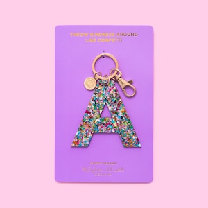 Może przedstawiać: Brelok do kluczy z literą A z kolorowych błyskotek, ze złotym kółkiem na klucze i małym złotym wisiorkiem. Brelok znajduje się na fioletowym tle z tekstem "Throw kindness around like confetti!" i "Confetti Keychain Taylor Elliott Designs".