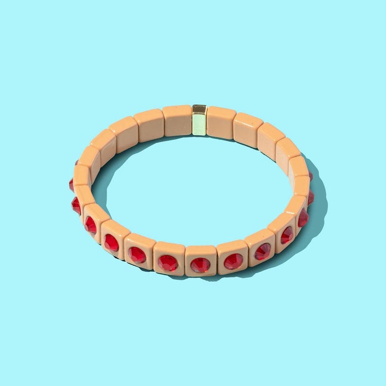 May include: A peach-colored bracelet with red gemstone accents. The bracelet is made of square beads and has a gold clasp.