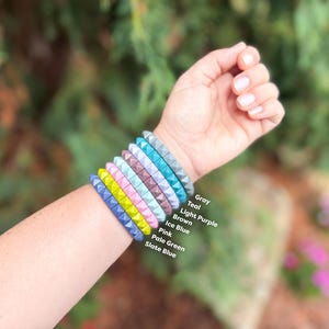 May include: A stack of seven silicone bracelets with a pyramid stud texture in slate blue, pale green, pink, ice blue, brown, light purple, teal, and gray. Each bracelet is labeled with its corresponding color name in white text next to the stack.