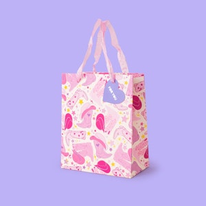 K&ouml;nnte beinhalten: Eine rosa Geschenkt&uuml;te mit wei&szlig;em Hintergrund und einem rosa-wei&szlig;en Muster aus Cowboystiefeln und Sternen. Die Tasche hat einen rosa Schleifenhenkel und ein lilafarbenes herzf&ouml;rmiges Etikett mit der Aufschrift "F&uuml;r dich".