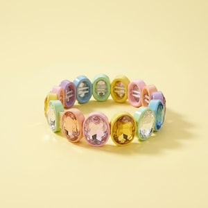 Op de afbeelding: Een pastelkleurige armband met ovale, edelsteenachtige kralen. De armband heeft een verscheidenheid aan kleuren, waaronder roze, geel, groen, blauw en paars. De edelstenen zijn gezet in bijpassende pastelkleurige zettingen.