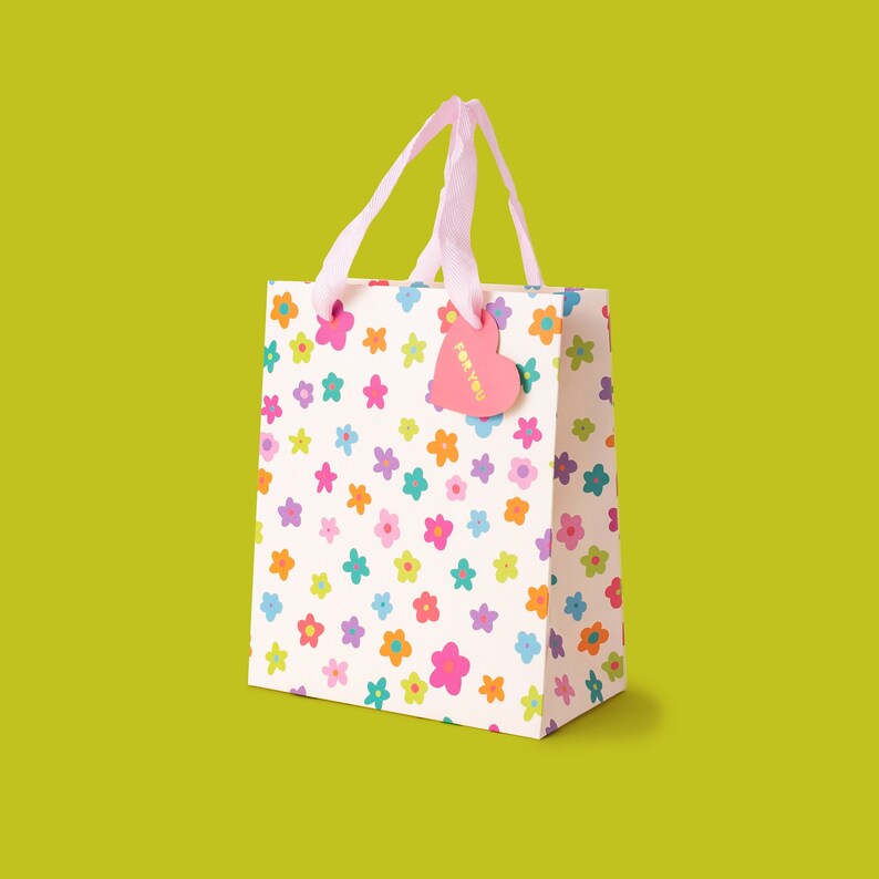 May include: A white gift bag with a pink handle and a pink heart-shaped tag that says "For You". The bag is covered in a pattern of colourful flowers.