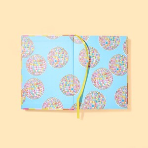 May include: Open notebook with a light blue cover featuring a pattern of colorful disco balls. The interior pages are not visible. A yellow ribbon bookmark is visible. The notebook is on a light orange background.