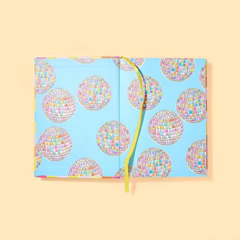May include: Open notebook with a light blue cover featuring a pattern of colorful disco balls. The interior pages are not visible. A yellow ribbon bookmark is visible. The notebook is on a light orange background.