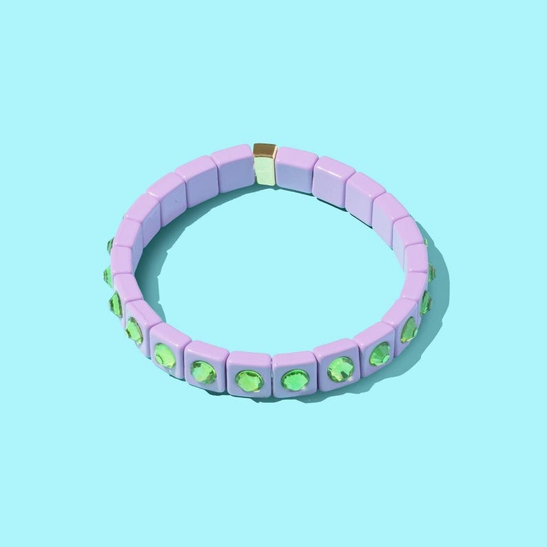 May include: A lilac-colored bracelet with green gemstone accents. The bracelet is made of square beads and has a gold clasp.