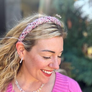 Può includere: Una fascia per capelli rosa e bianca lavorata a maglia è indossata da una persona con i capelli biondi. La fascia è il punto focale, con un aspetto strutturato. La persona indossa un top rosa e gioielli. Il nome "Julia" è scritto sull'immagine.