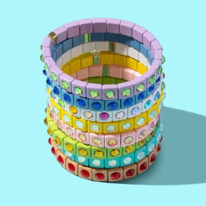 Op de afbeelding: Een stapel kleurrijke armbanden gemaakt van vierkante kralen met strass. De armbanden zijn in tinten paars, blauw, geel, groen en rood.